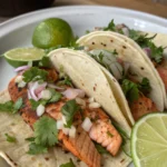 Grilled salmon fish tacos with fresh cabbage slaw, creamy sauce, and lime wedges.